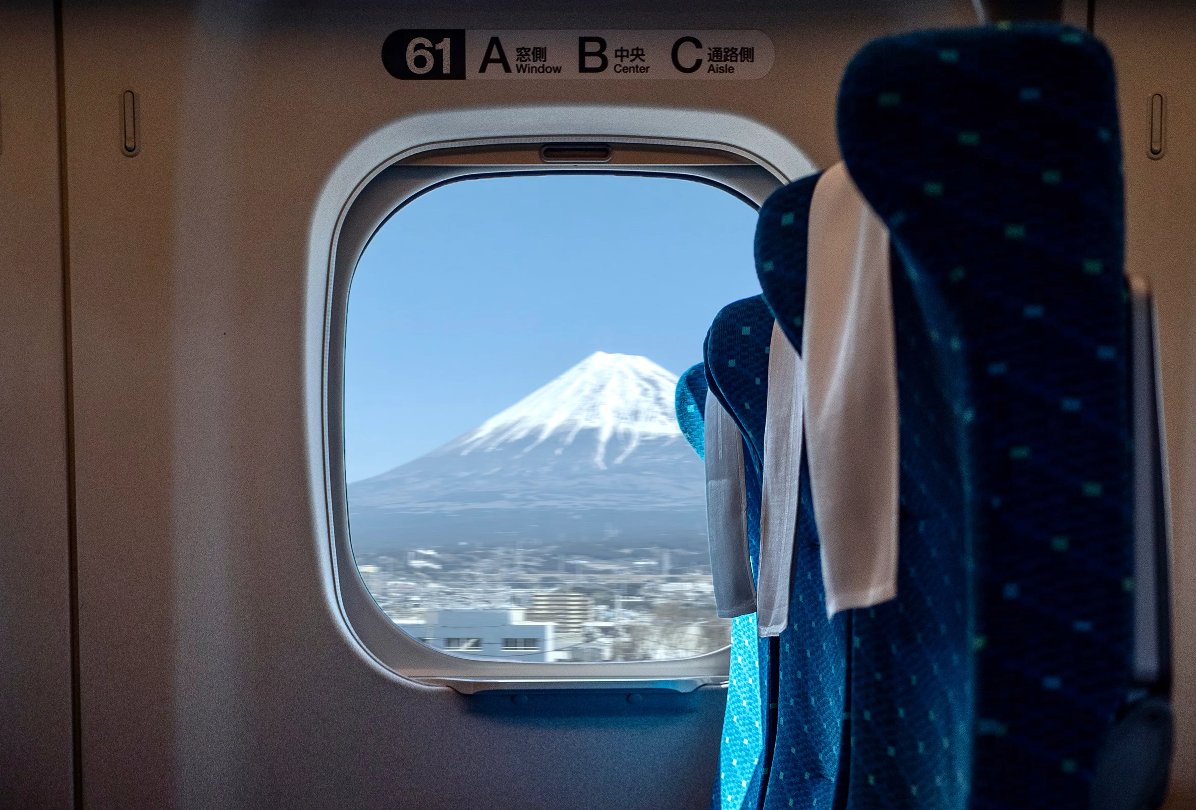 Interior view of a train carriage with a three-seat row, framed by a window view of Mount Fuji against the sky.
